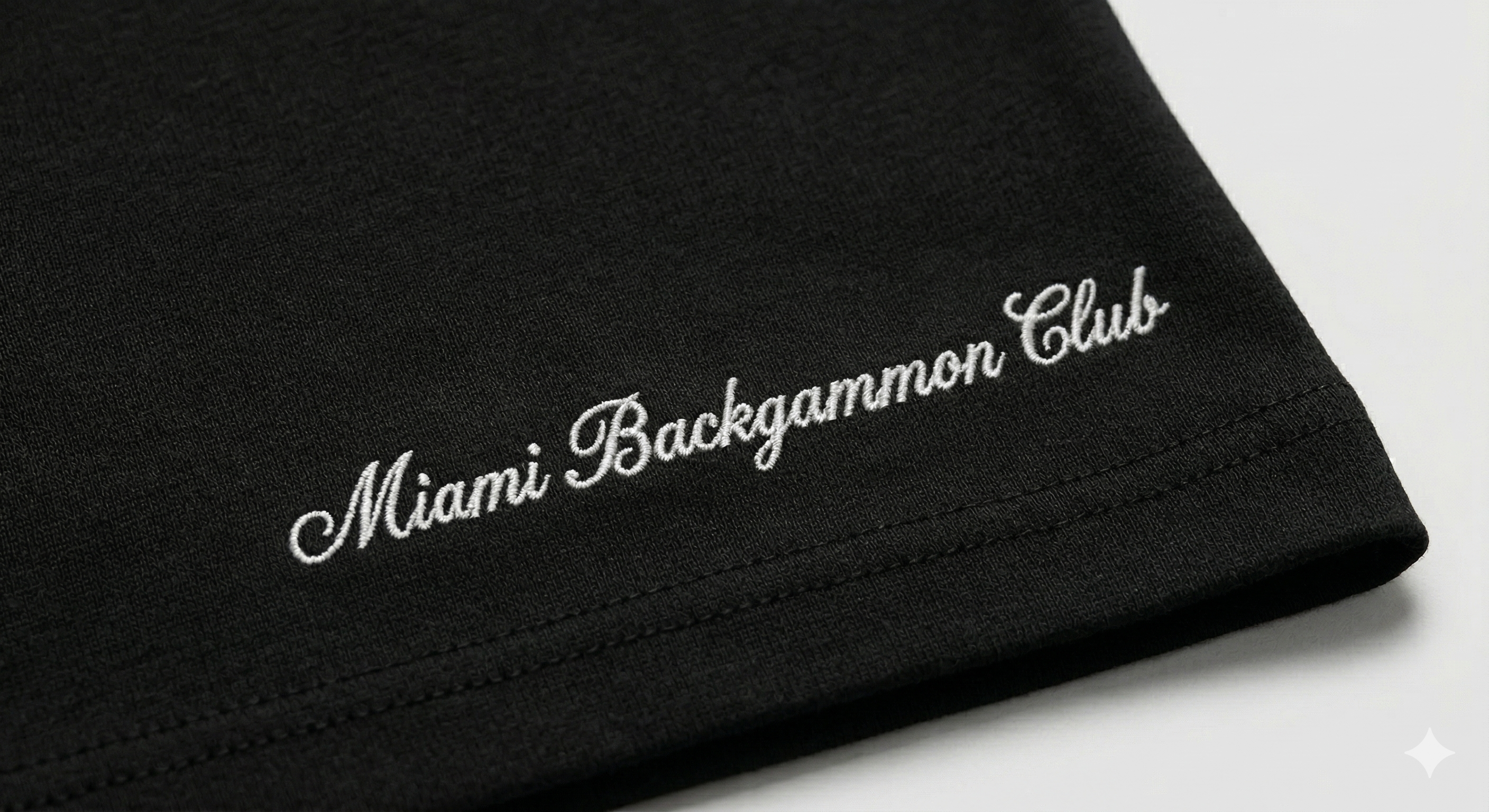 Close-up of the Miami Backgammon Club script logo embroidered on the hem of the black Signature Script Tee