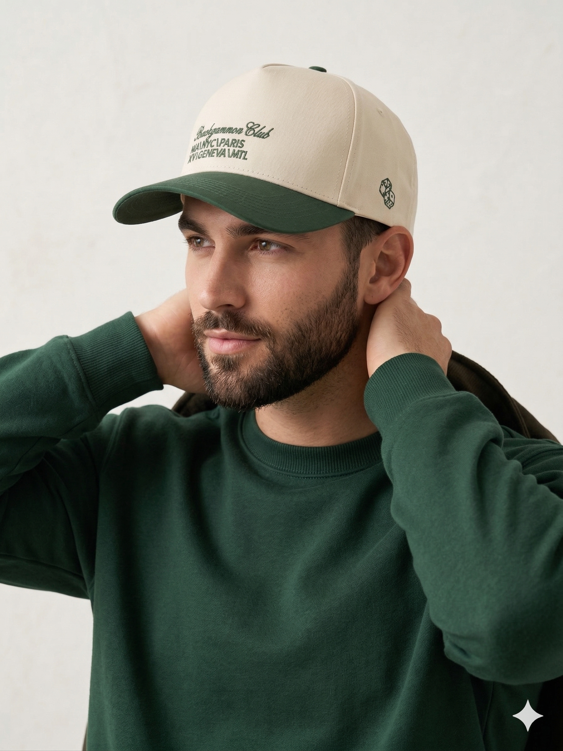 Model wearing the Backgammon Club Signature Trucker Hat with cream crown, forest green brim, and embroidered city list, paired with a green sweatshirt