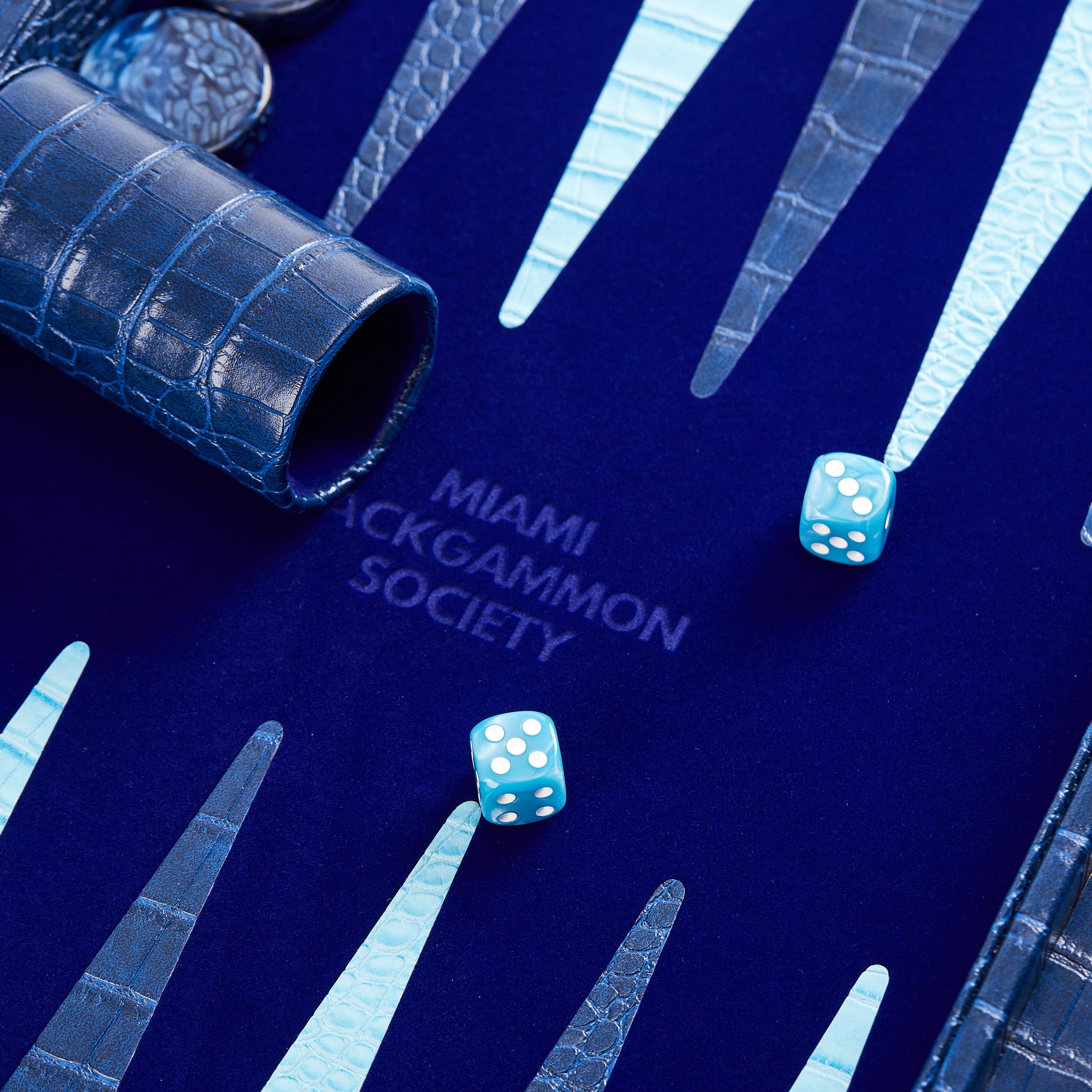 Close-up of dice cup and cyan dice resting on the Midnight Navy playing field with Miami Backgammon Society logo embossed at center