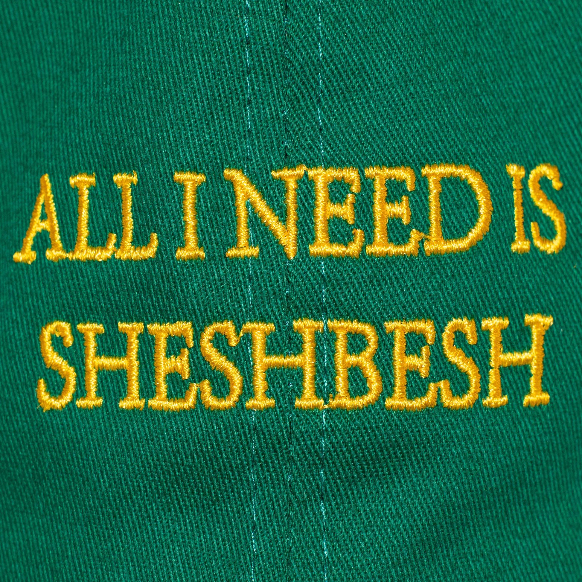Macro close-up of the embroidered 'All I Need Is SheshBesh' text in yellow thread on the green cap