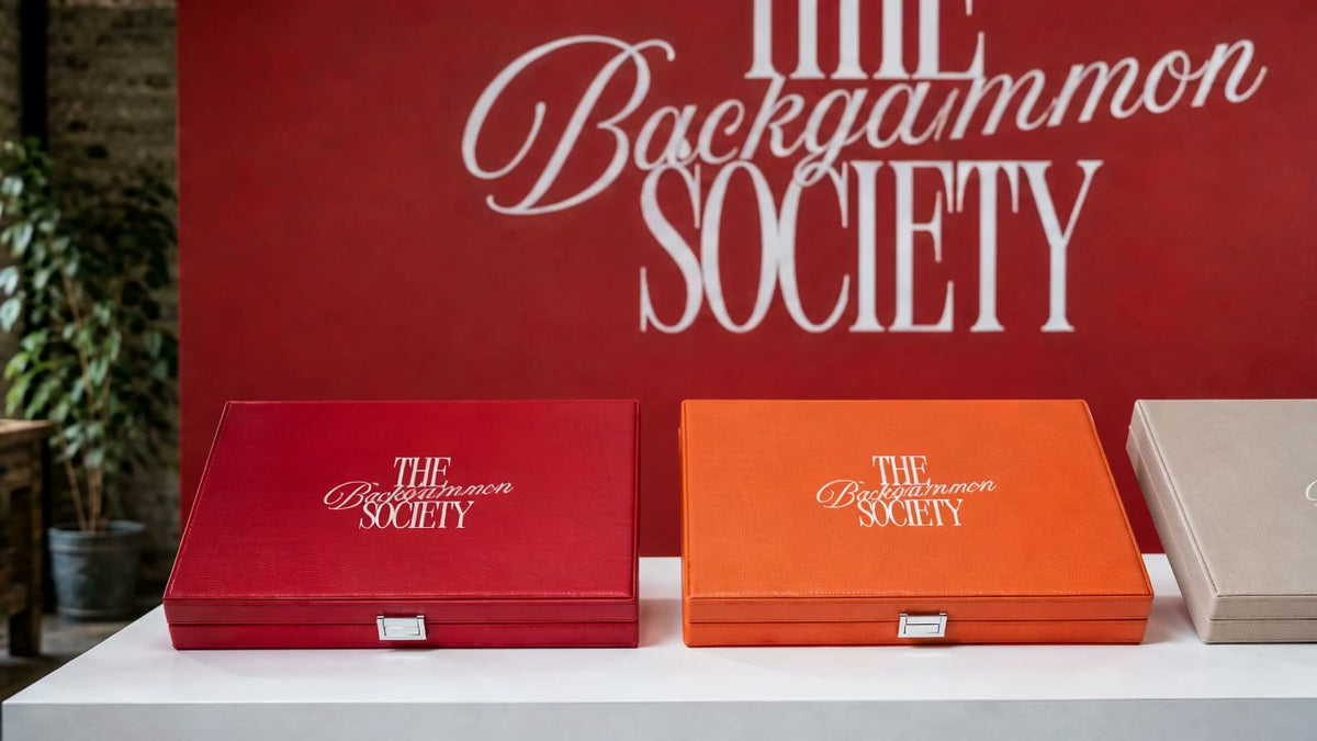 Trois mallettes The Backgammon Society alignees sur une console devant un mur rouge au logo de la marque